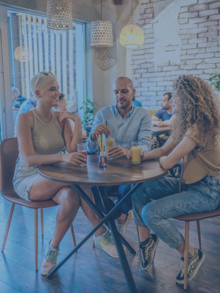 Three adults sitting at a public table chatting and drinking what appears to be iced coffees.