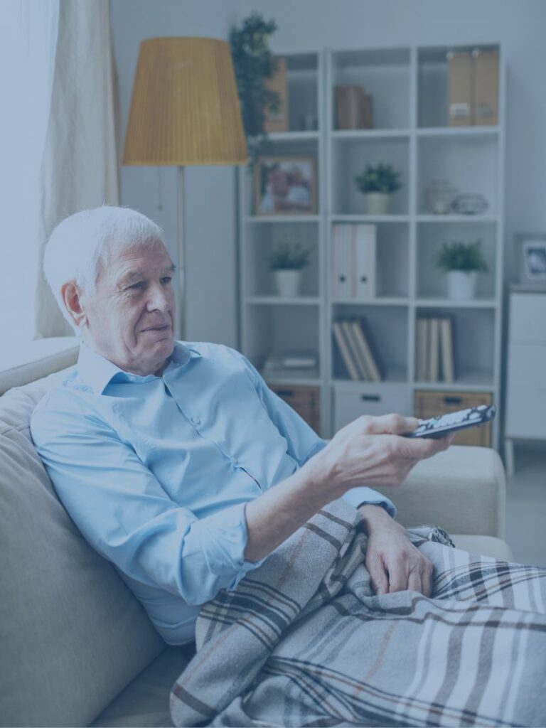 Older gentleman sitting on a couch with a blanket holding a TV remote.